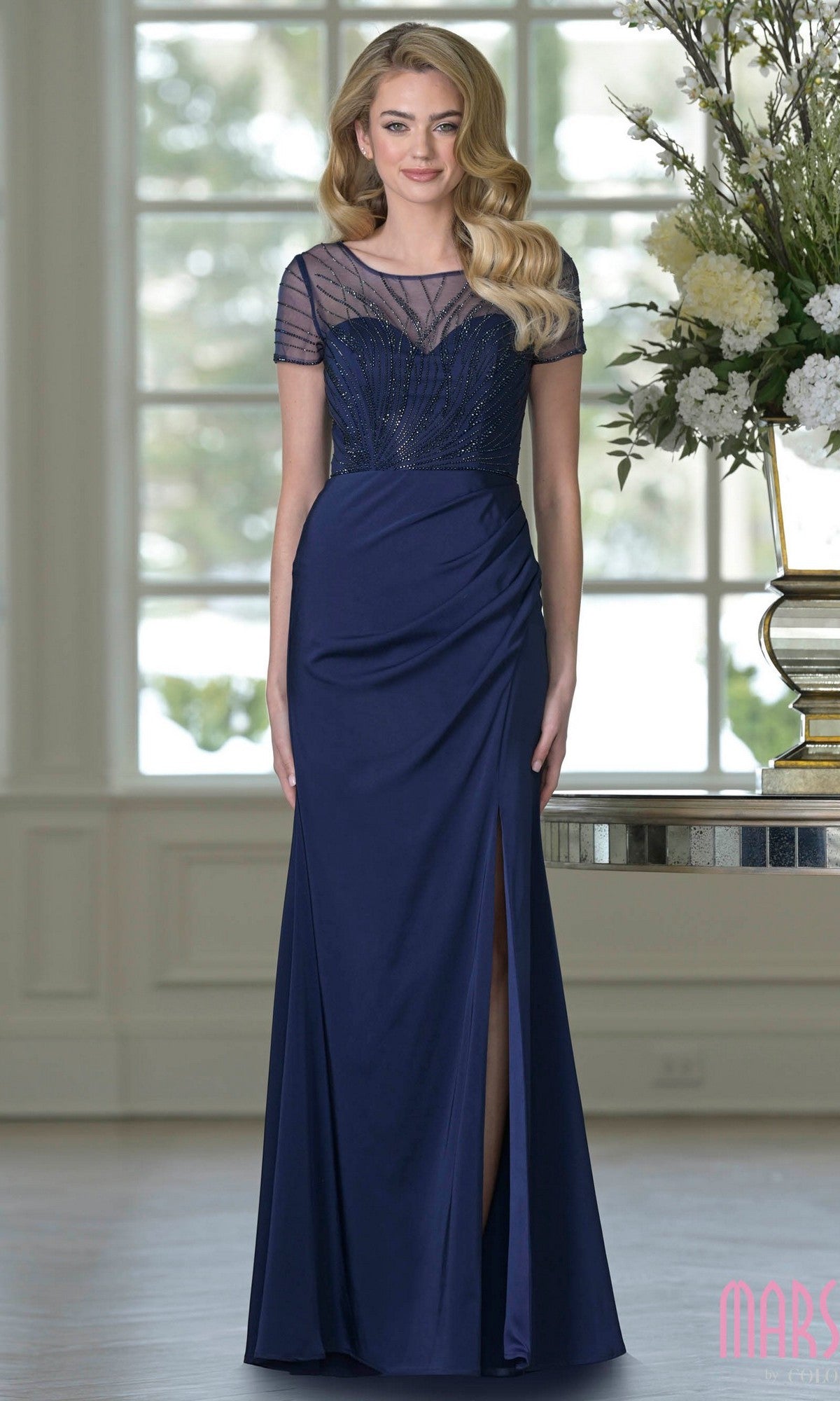 A woman stands indoors by a flower-adorned table, wearing the Marsoni M350 long formal dress in navy with a beaded bodice, short sheer sleeves, ruched waist, and a sheath skirt featuring a front slit.